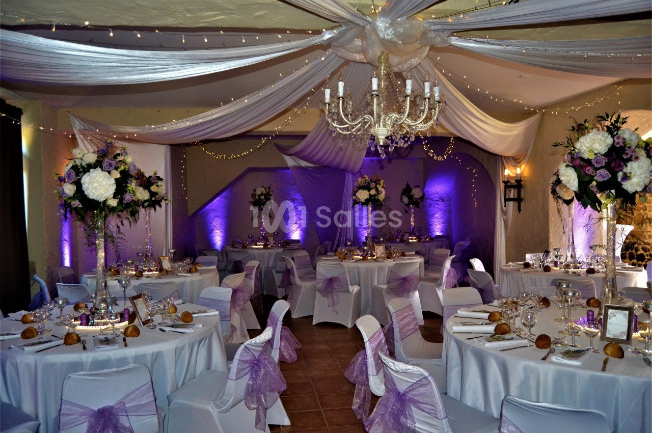 Salle de réception décorée avec des nappes blanches, des rubans violets, des fleurs et un éclairage tamisé.
