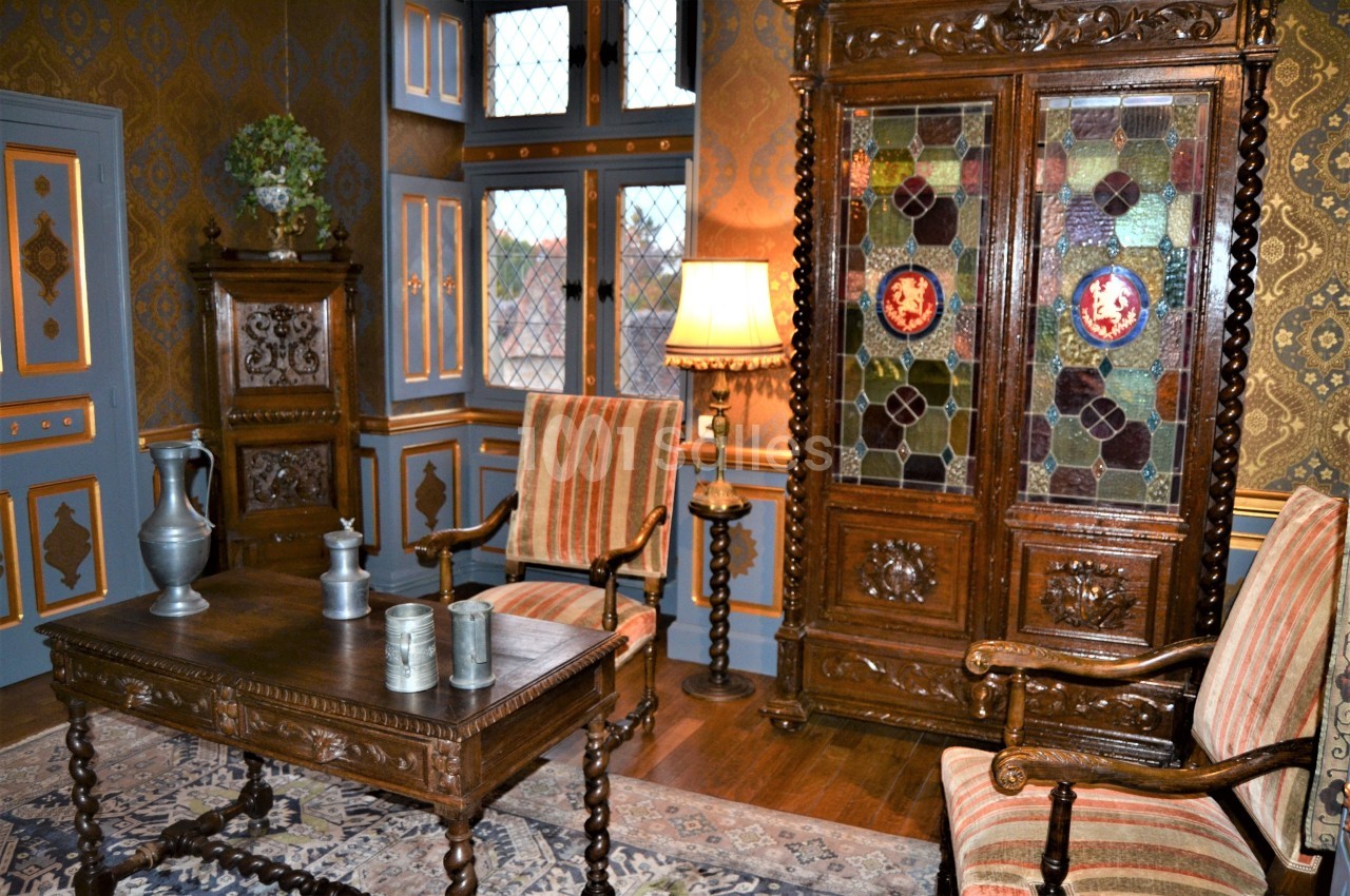 Salle de style ancien avec mobilier en bois sculpté, vitrail coloré, tapis et chaises rembourrées à rayures.