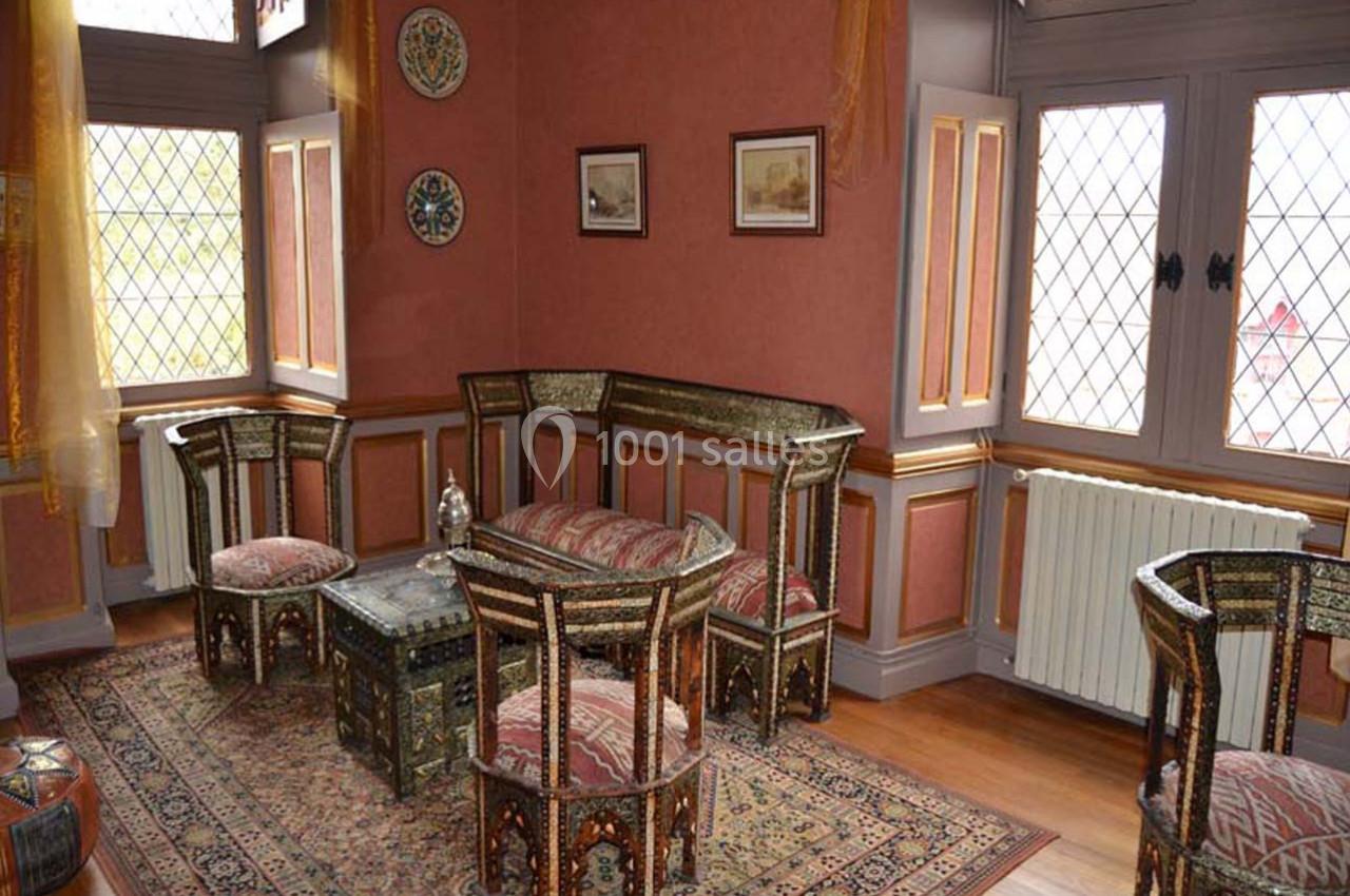 Salon décoré dans un style oriental avec mobilier en bois incrusté, tapis coloré et fenêtres à vitraux.