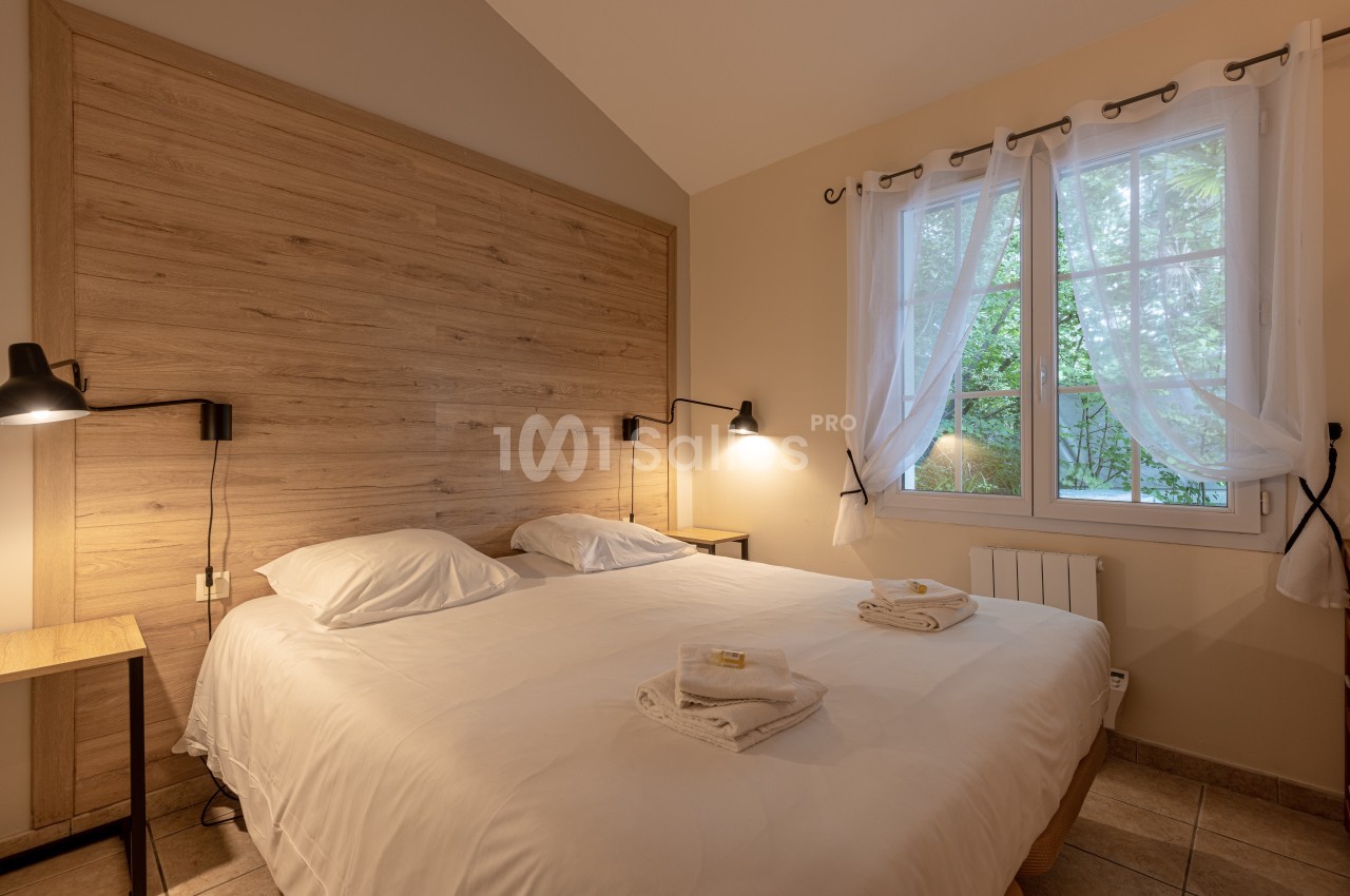 Chambre avec lit double, tête de lit en bois, lampes murales et fenêtre avec rideaux donnant sur un jardin.