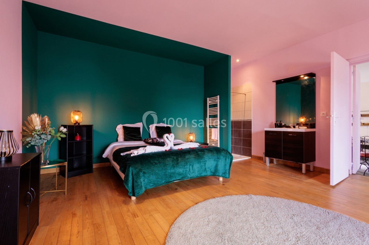 Chambre lumineuse avec lit double, mur vert, parquet, tapis rond et salle de bain attenante visible.