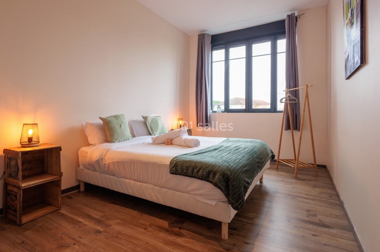 Chambre lumineuse avec lit double, parquet, table de chevet, portant à vêtements et grande fenêtre avec rideaux.