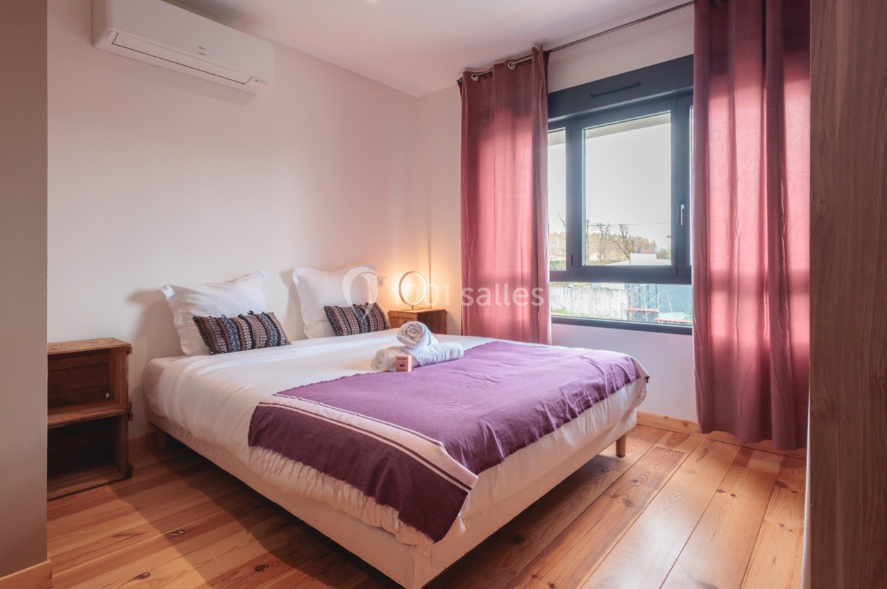 Chambre lumineuse avec lit double, draps violets, parquet en bois, fenêtre avec rideaux et climatisation murale.