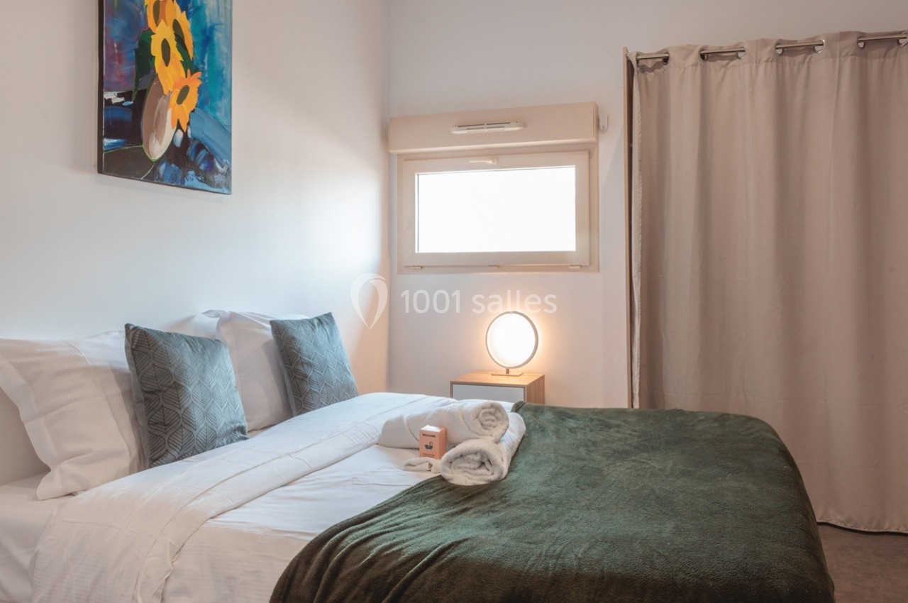 Chambre lumineuse avec un lit double, des coussins gris, une couverture verte, une lampe de chevet et un tableau au mur.