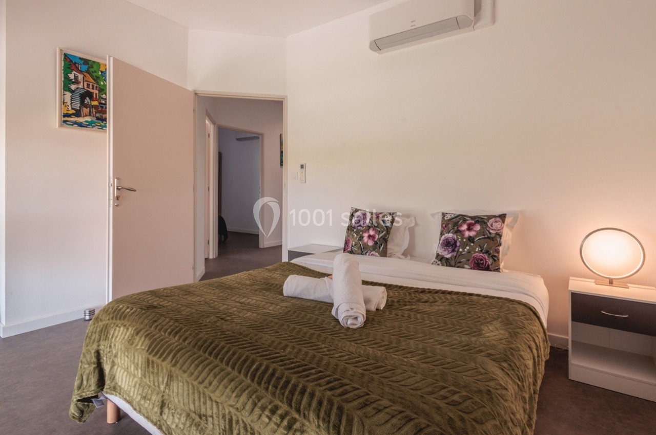 Chambre lumineuse avec lit double, couvre-lit vert, coussins fleuris, table de chevet et porte ouverte sur un couloir.