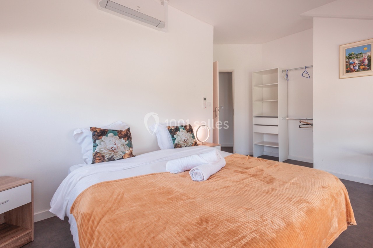 Chambre lumineuse avec lit double, couverture orange, coussins fleuris, placard ouvert et climatisation murale.