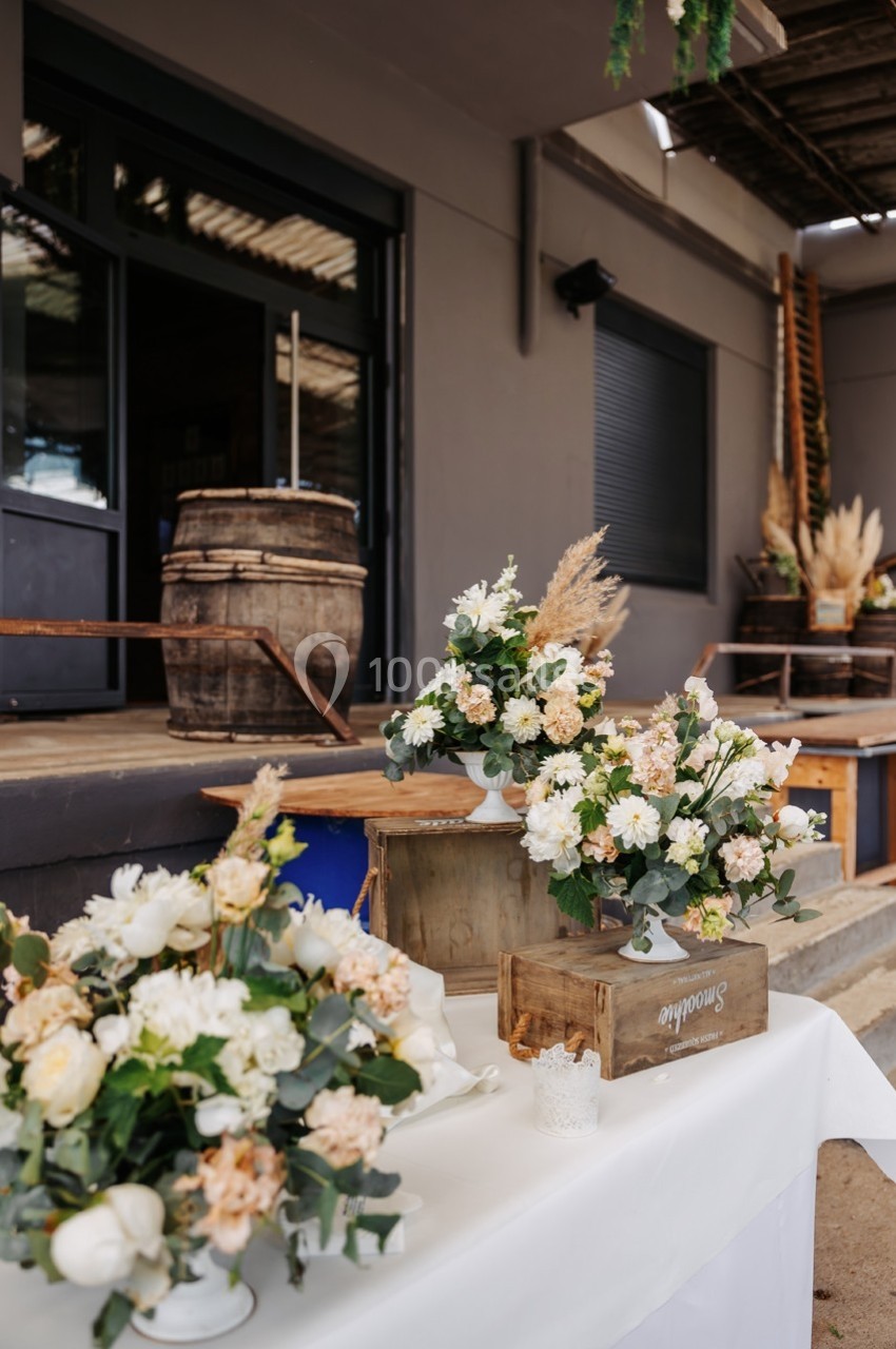 Arrangements floraux blancs et beiges sur une table en extérieur, près de caisses en bois et d'un bâtiment industriel.