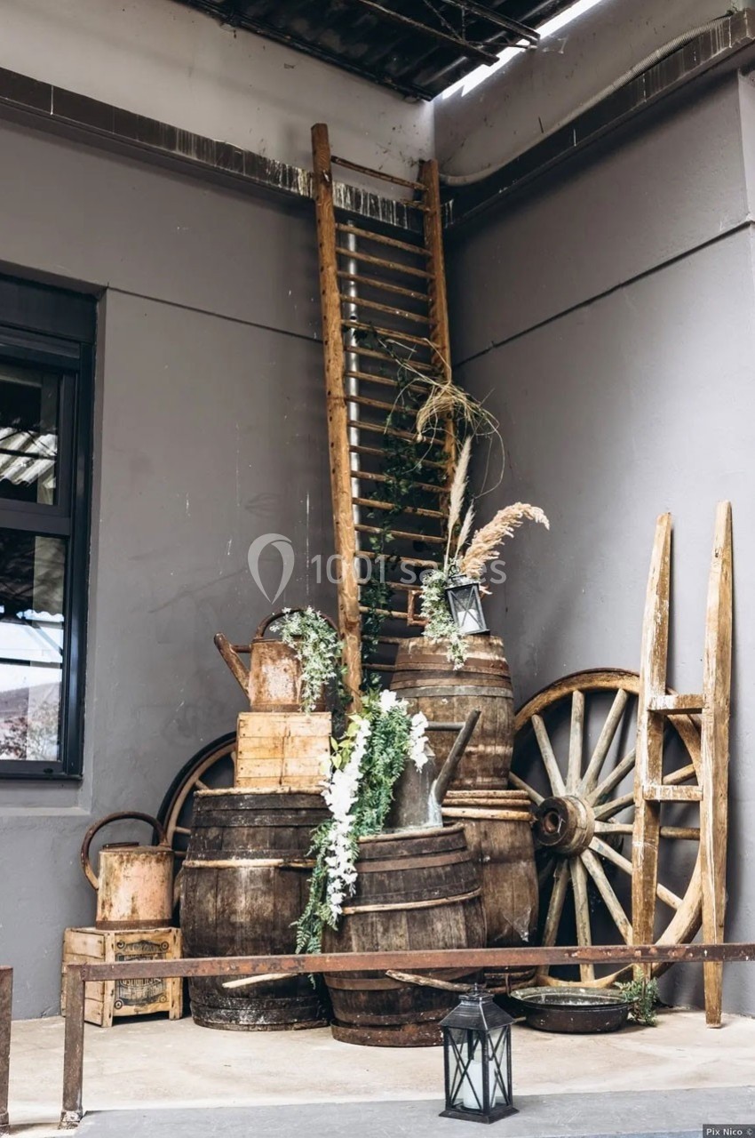 Décoration rustique avec tonneaux en bois, échelle, roues de charrette et plantes dans un coin de mur gris.
