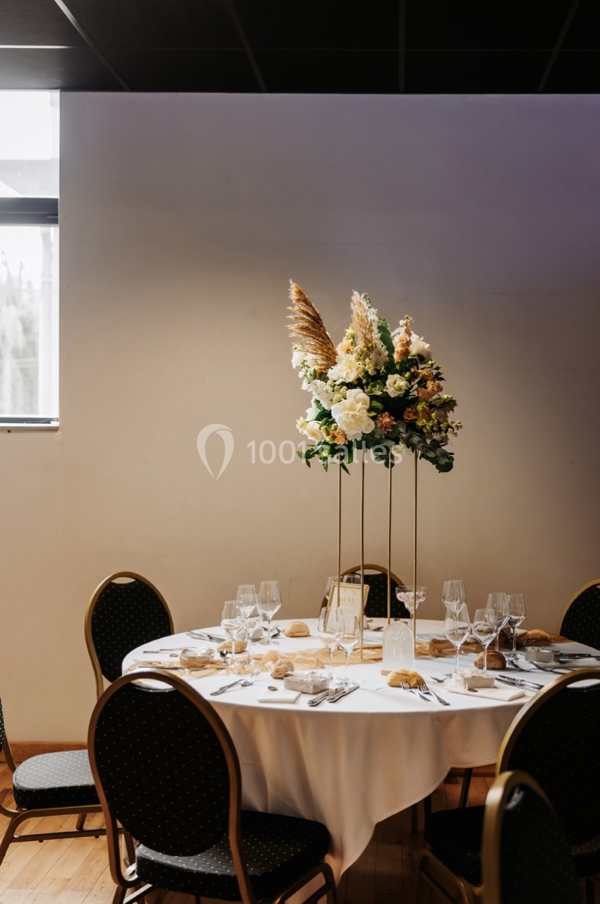 Table ronde élégamment dressée avec nappes blanches, verres, couverts et un centre de table floral surélevé.