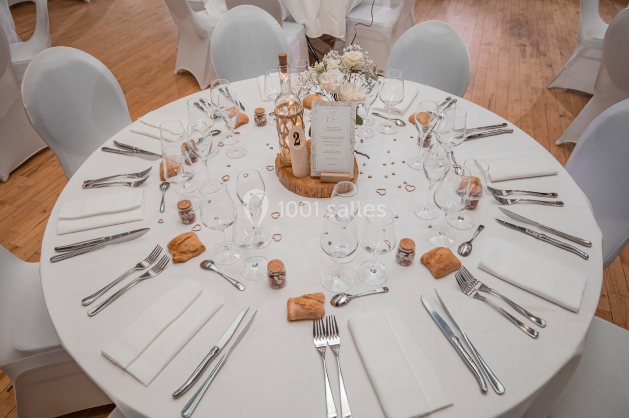 Table ronde élégamment dressée avec nappes blanches, couverts, verres, menu central et décorations florales.