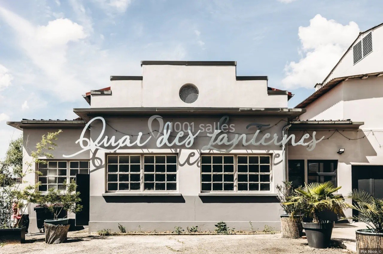 Façade d'un bâtiment gris avec de grandes fenêtres, une inscription ’Quai des Lanternes’ et des plantes en pots devant.
