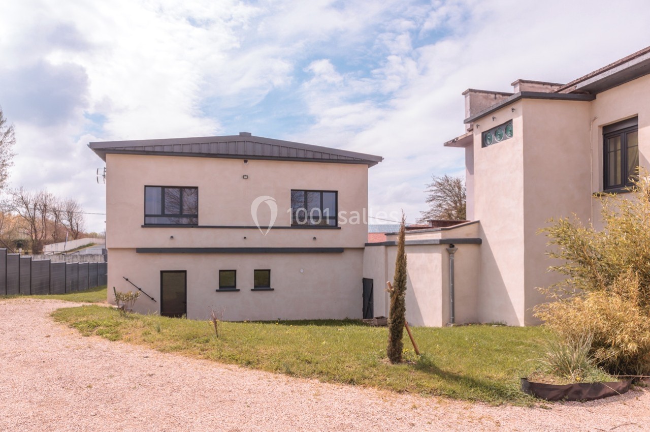 Bâtiment moderne avec façade beige, fenêtres noires et jardin herbeux, sous un ciel partiellement nuageux.