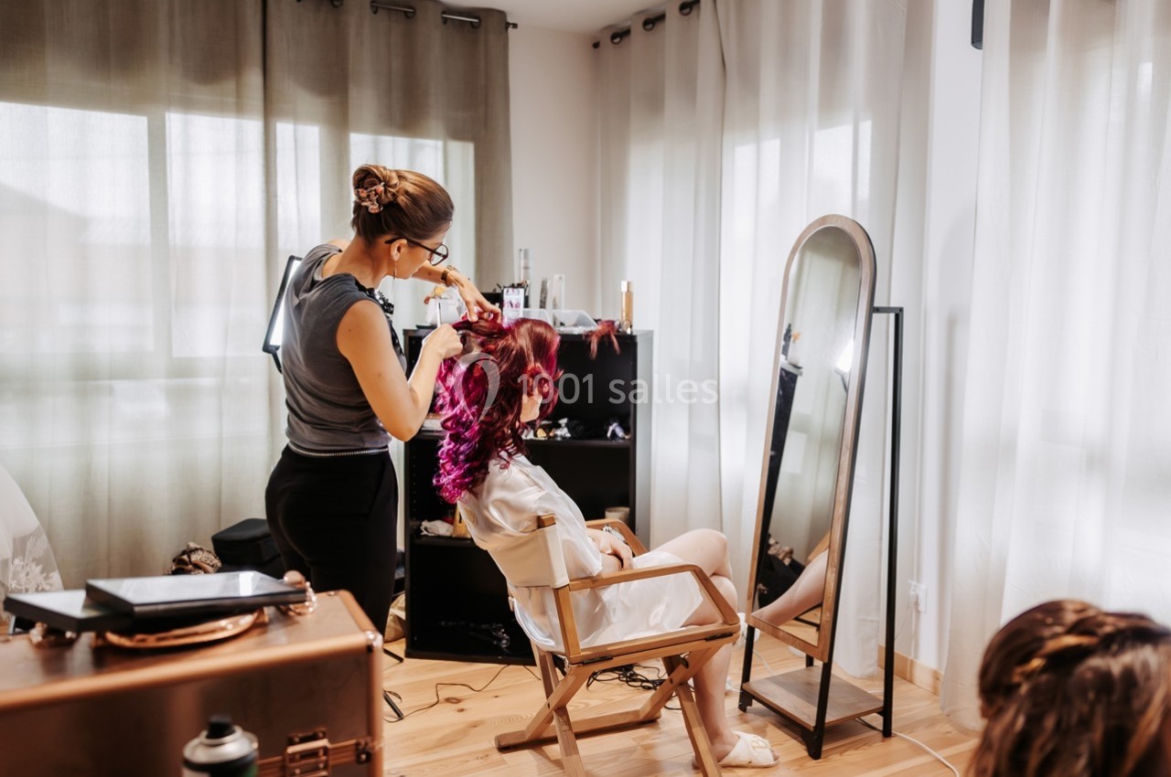 Une coiffeuse stylise les cheveux d'une femme assise devant un miroir dans une pièce lumineuse.