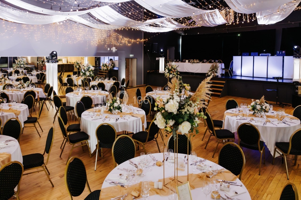 Salle de réception décorée avec des tables rondes, nappes blanches, chaises noires et arrangements floraux élégants.