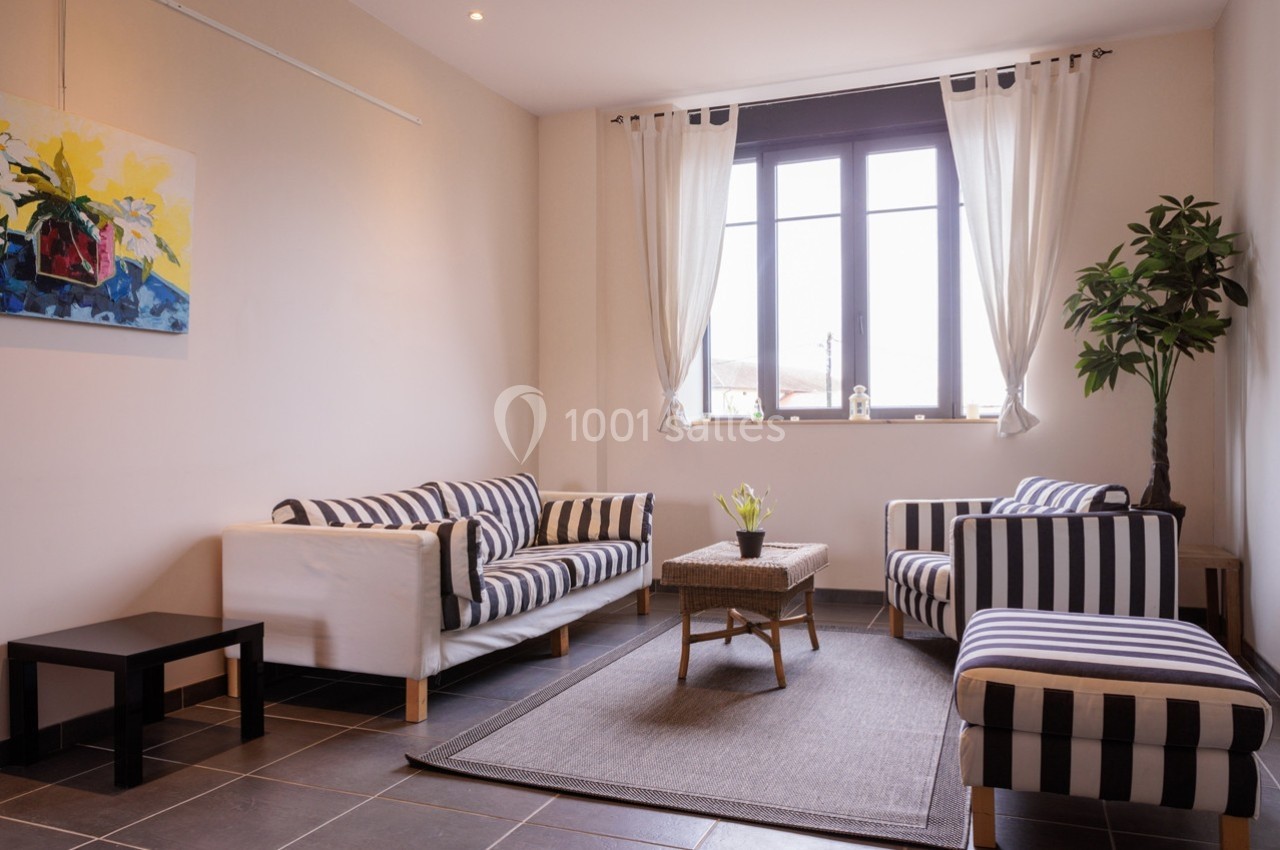 Salon lumineux avec canapé et fauteuils rayés, table basse en rotin, plante verte et grande fenêtre avec rideaux blancs.