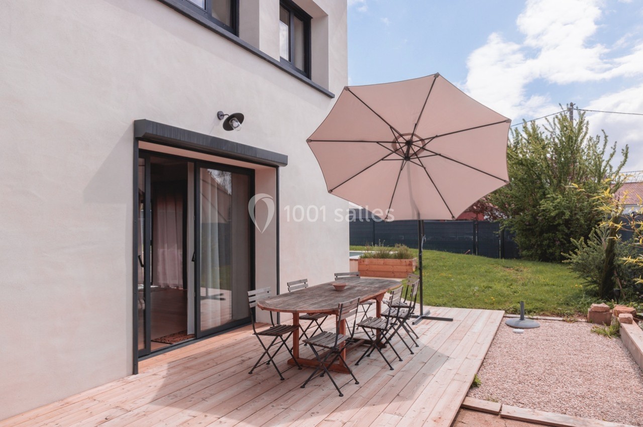 Terrasse en bois avec table, chaises et parasol beige, jouxtant une maison moderne et un jardin verdoyant.