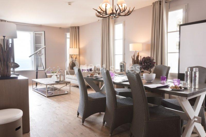Salle à manger lumineuse avec table en bois, chaises en osier, canapé beige et grandes fenêtres laissant entrer la lumière.