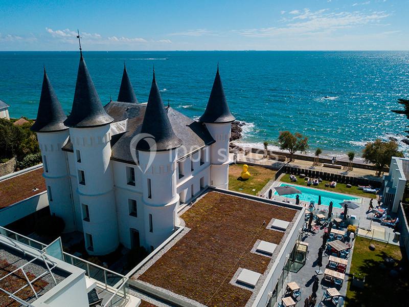 Vue aérienne d'un château blanc avec des tours, situé en bord de mer, entouré d'une piscine et d'espaces verts.