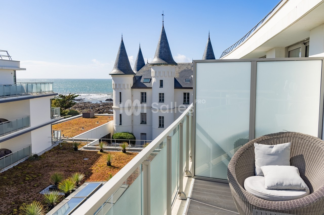 Vue depuis un balcon moderne avec fauteuil, donnant sur un château en bord de mer et des bâtiments contemporains.