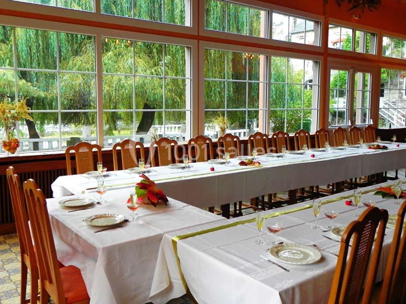 Salle lumineuse avec de grandes baies vitrées, tables dressées avec nappes blanches et chaises en bois.