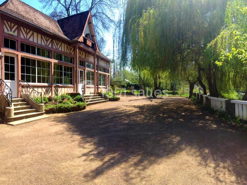 Bâtiment à colombages avec grandes fenêtres, entouré d'arbres et d'un chemin ombragé dans un cadre naturel.