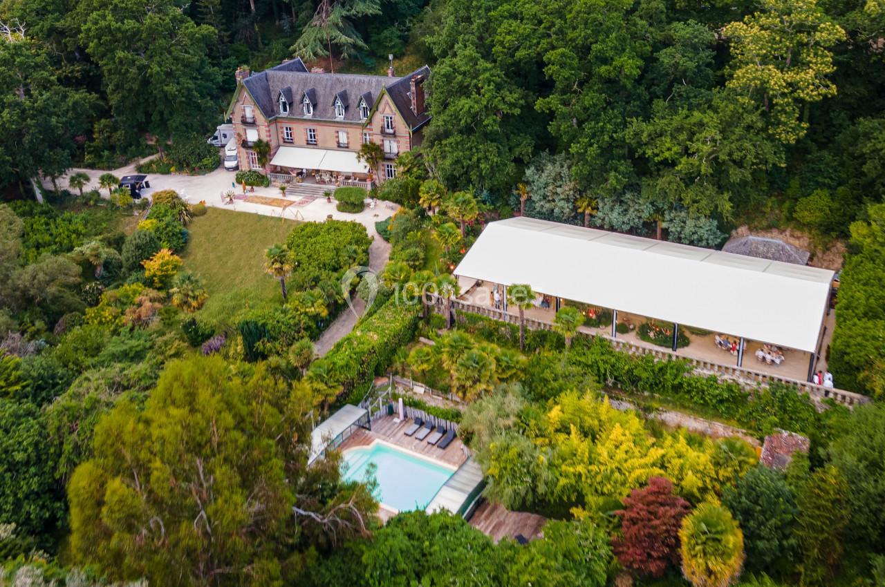 Vue aérienne d'une grande maison entourée de jardins verdoyants, avec une piscine et une terrasse couverte.