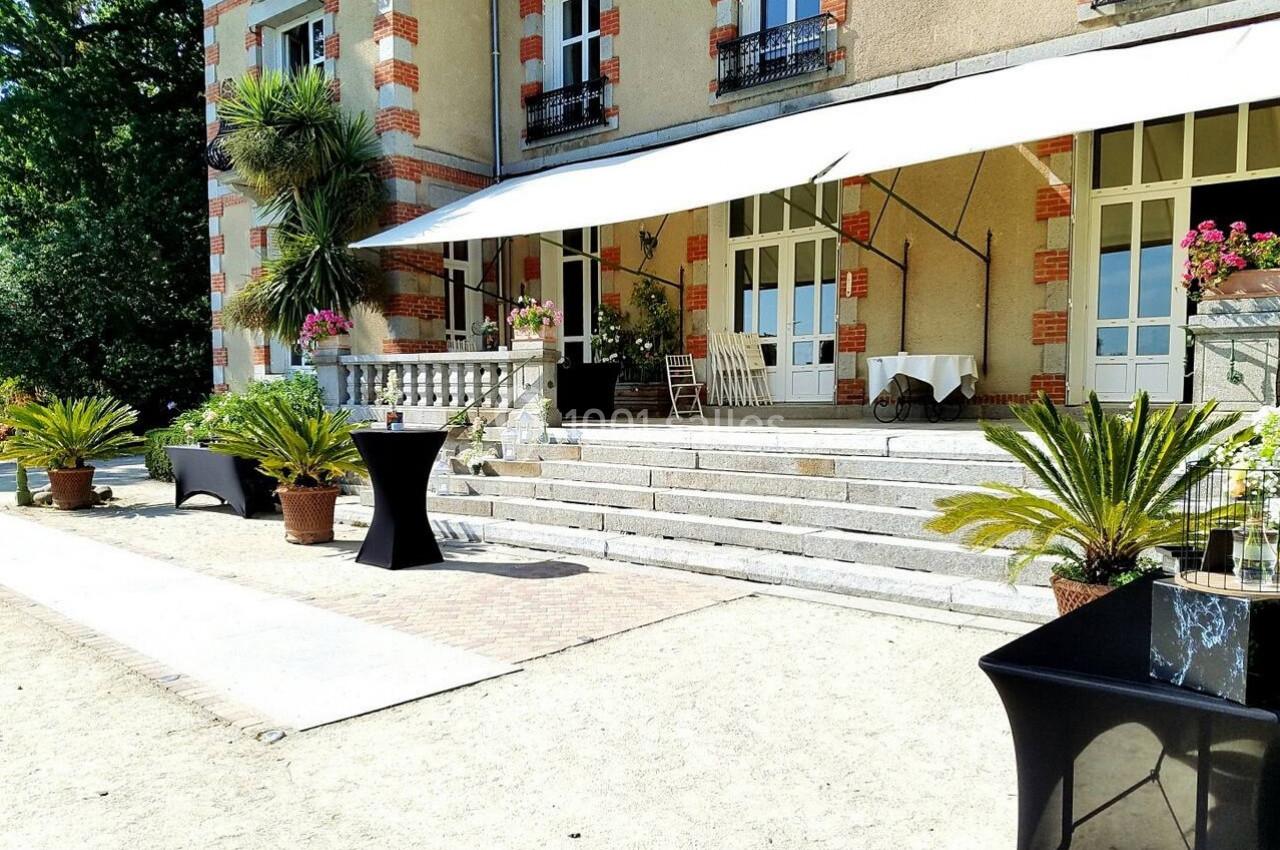 Terrasse ensoleillée d'un bâtiment ancien avec des tables hautes noires, des plantes en pots et un auvent blanc.