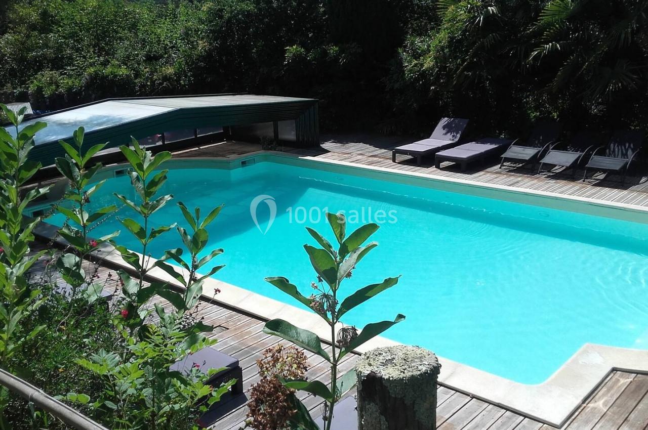 Piscine extérieure entourée de végétation, avec des transats et un abri partiellement ouvert sur le côté.