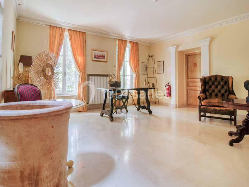 Salle lumineuse avec mobilier ancien, fauteuils, bureau et grandes fenêtres encadrées de rideaux orange.