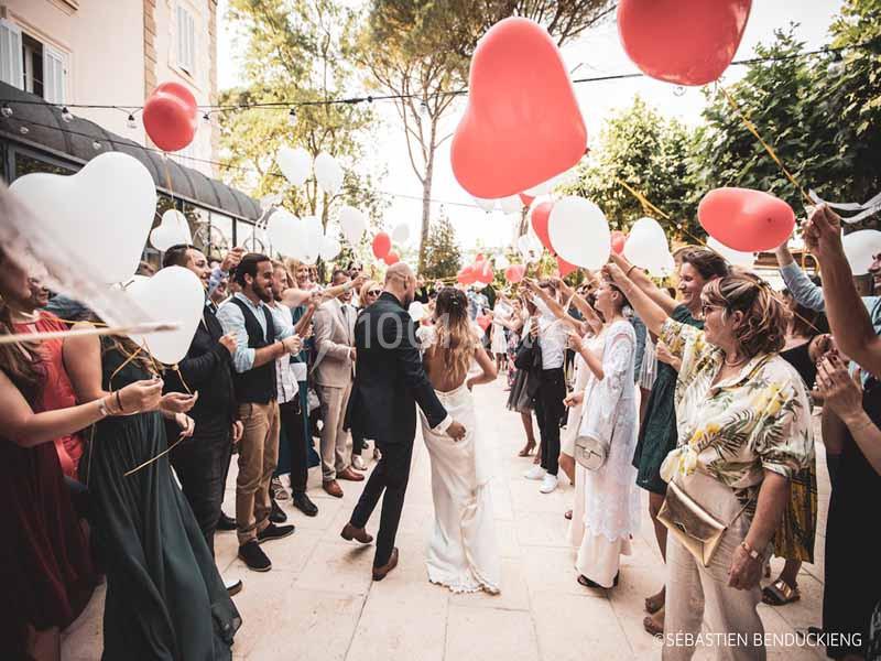 Un couple de mariés marche entre des invités tenant des ballons rouges et blancs dans une allée extérieure festive.