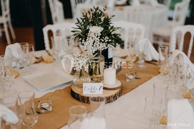 Centre de table avec fleurs blanches, feuillage, bougie et numéro de table, disposé sur une nappe beige.