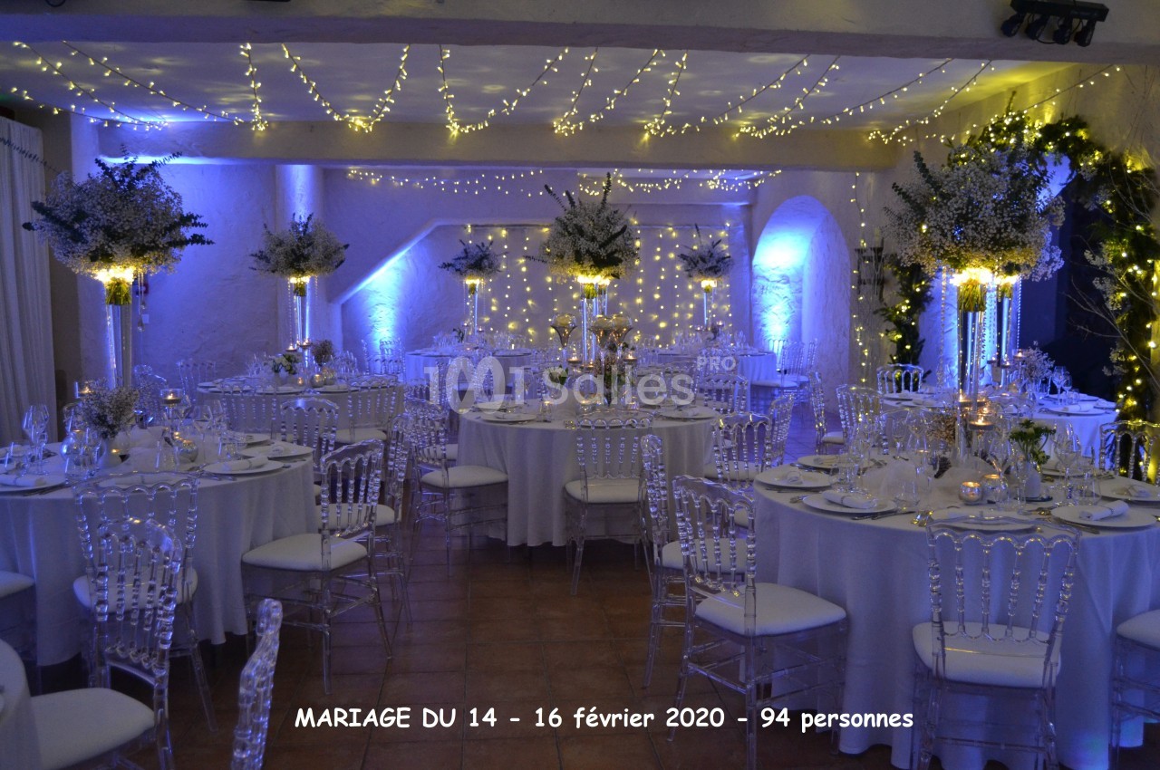 Salle de réception décorée pour un mariage avec tables rondes, chaises transparentes et éclairage chaleureux.