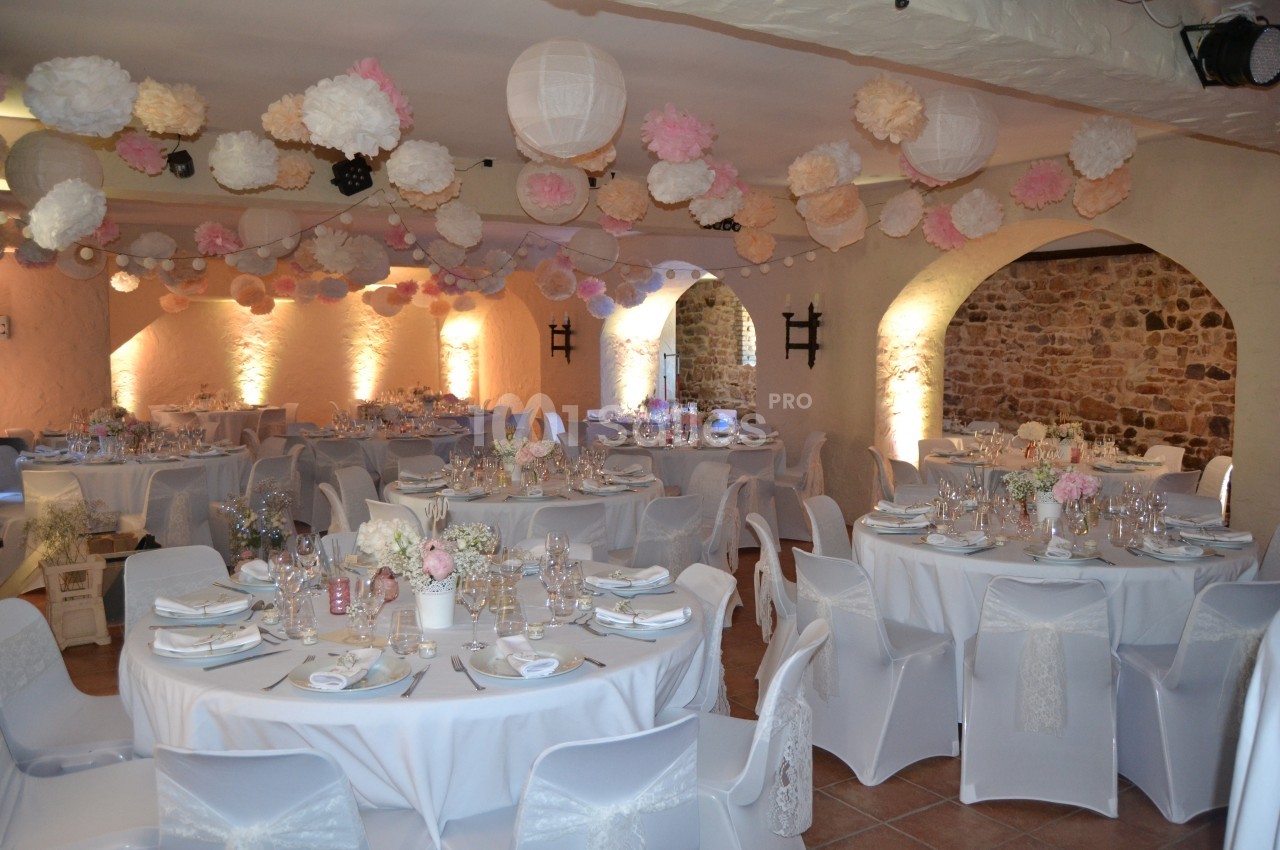 Salle de réception décorée avec des tables rondes, nappes blanches, vaisselle élégante et guirlandes de lanternes suspendues.