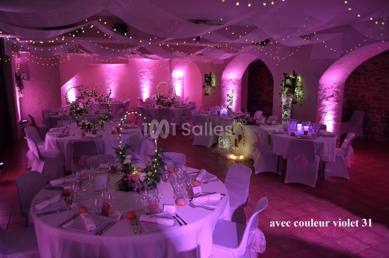 Salle de réception décorée avec des tables rondes, nappes blanches, éclairage violet et guirlandes lumineuses suspendues.