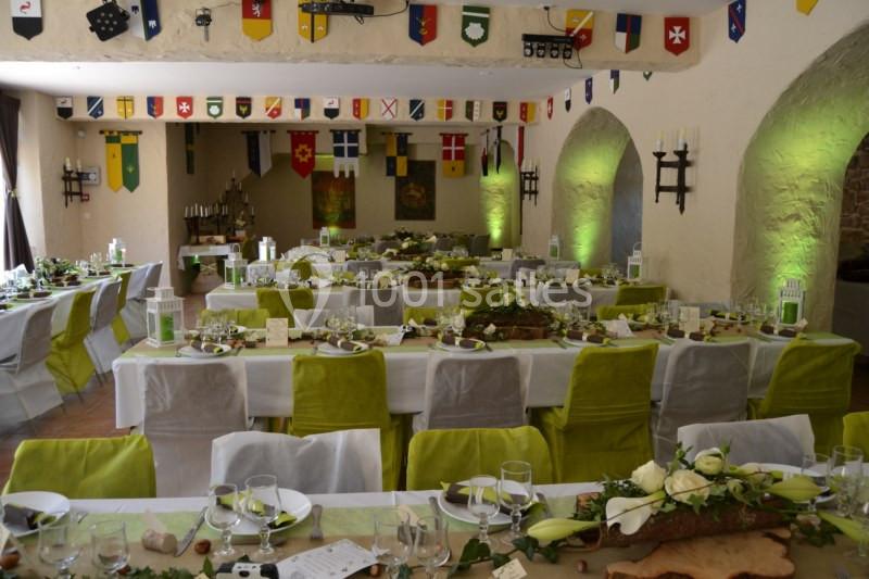 Salle décorée pour un banquet avec des tables alignées, nappes blanches, chaises vertes et décorations médiévales.