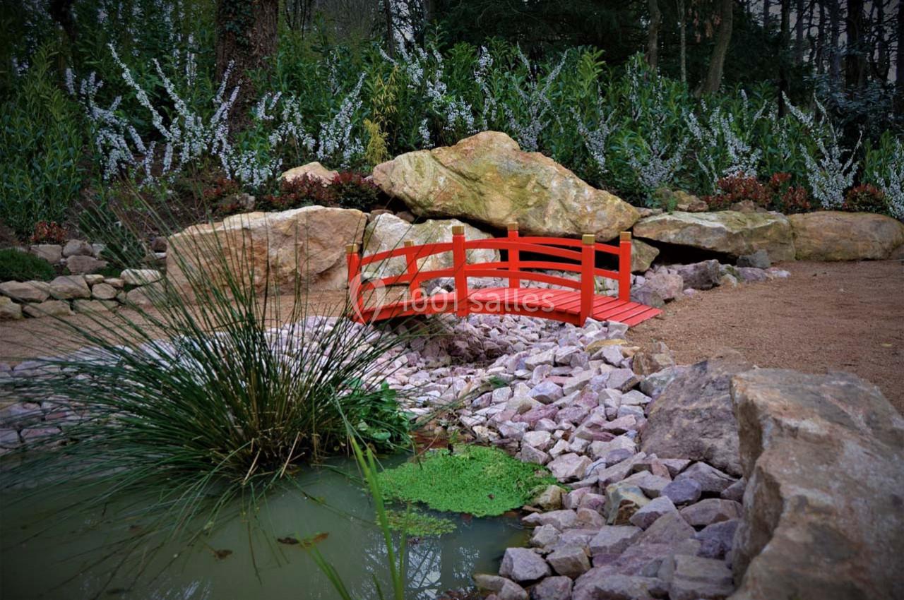 Petit pont rouge en bois traversant un ruisseau entouré de pierres, de végétation et d'un étang avec des plantes aquatiques.