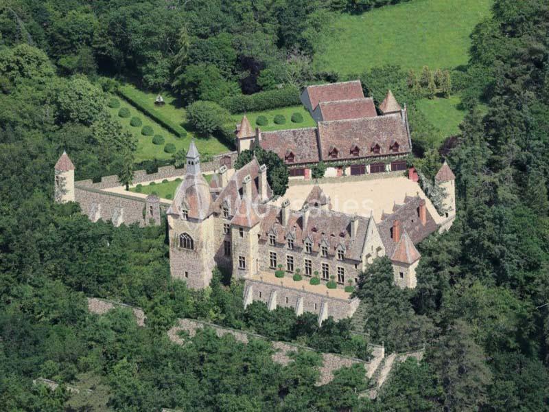 Vue aérienne d'un château médiéval entouré de jardins, de murs fortifiés et de forêt dense.