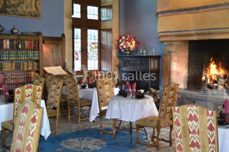 Salle chaleureuse avec cheminée allumée, tables dressées, chaises tapissées et bibliothèque en arrière-plan.
