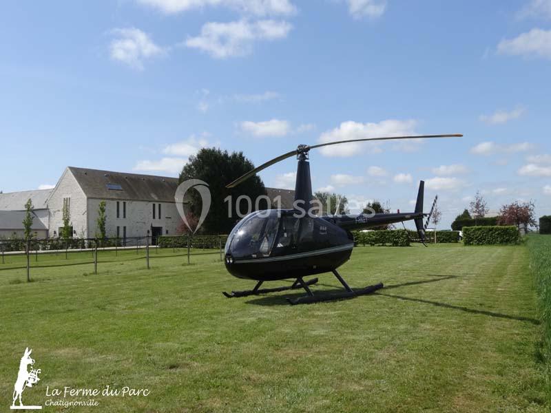 Hélicoptère noir posé sur une pelouse, avec des bâtiments et des arbres en arrière-plan sous un ciel bleu.
