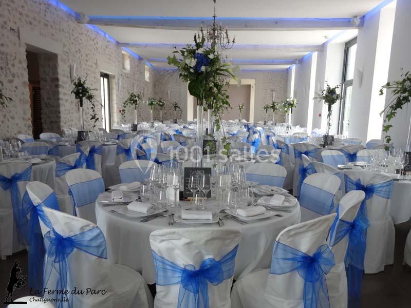 Salle de réception décorée avec des tables rondes, nappes blanches, chaises ornées de rubans bleus et centres de table…