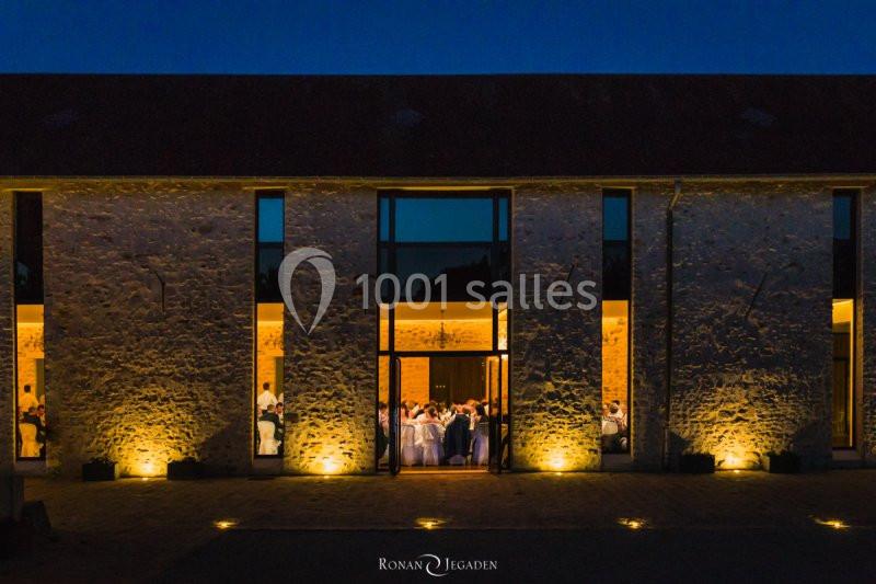 Façade en pierre éclairée d'un bâtiment avec de grandes fenêtres laissant entrevoir un événement en soirée.