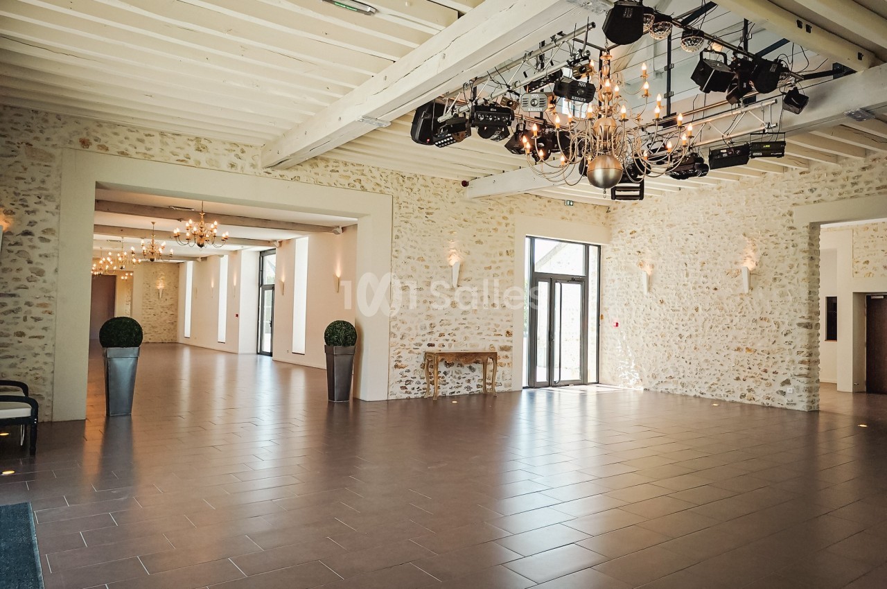 Salle spacieuse avec murs en pierre, grandes baies vitrées, poutres apparentes et éclairage suspendu moderne.
