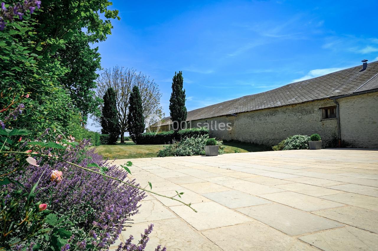 Cour pavée bordée de fleurs et d'arbustes, avec un bâtiment en pierre et des arbres sous un ciel bleu dégagé.