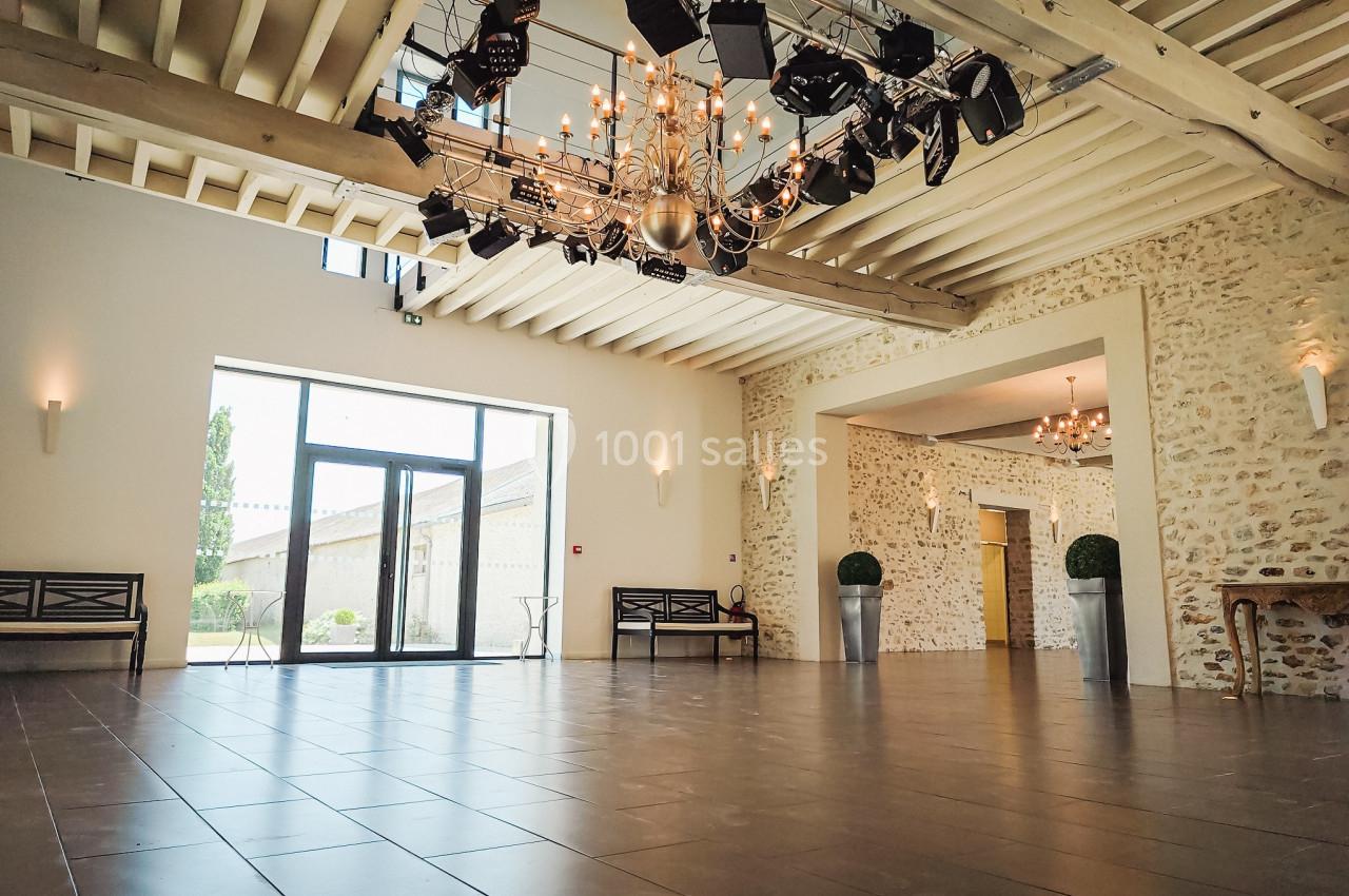 Salle spacieuse avec murs en pierre, sol carrelé, grandes baies vitrées et lustres suspendus au plafond en poutres…