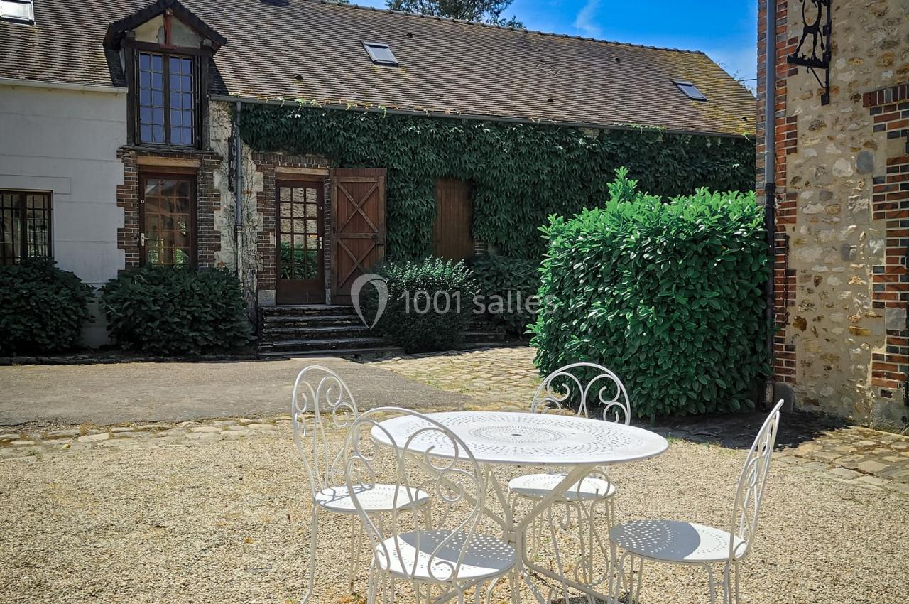 Table et chaises blanches en fer forgé sur une cour en gravier, devant des bâtiments en pierre et végétation.