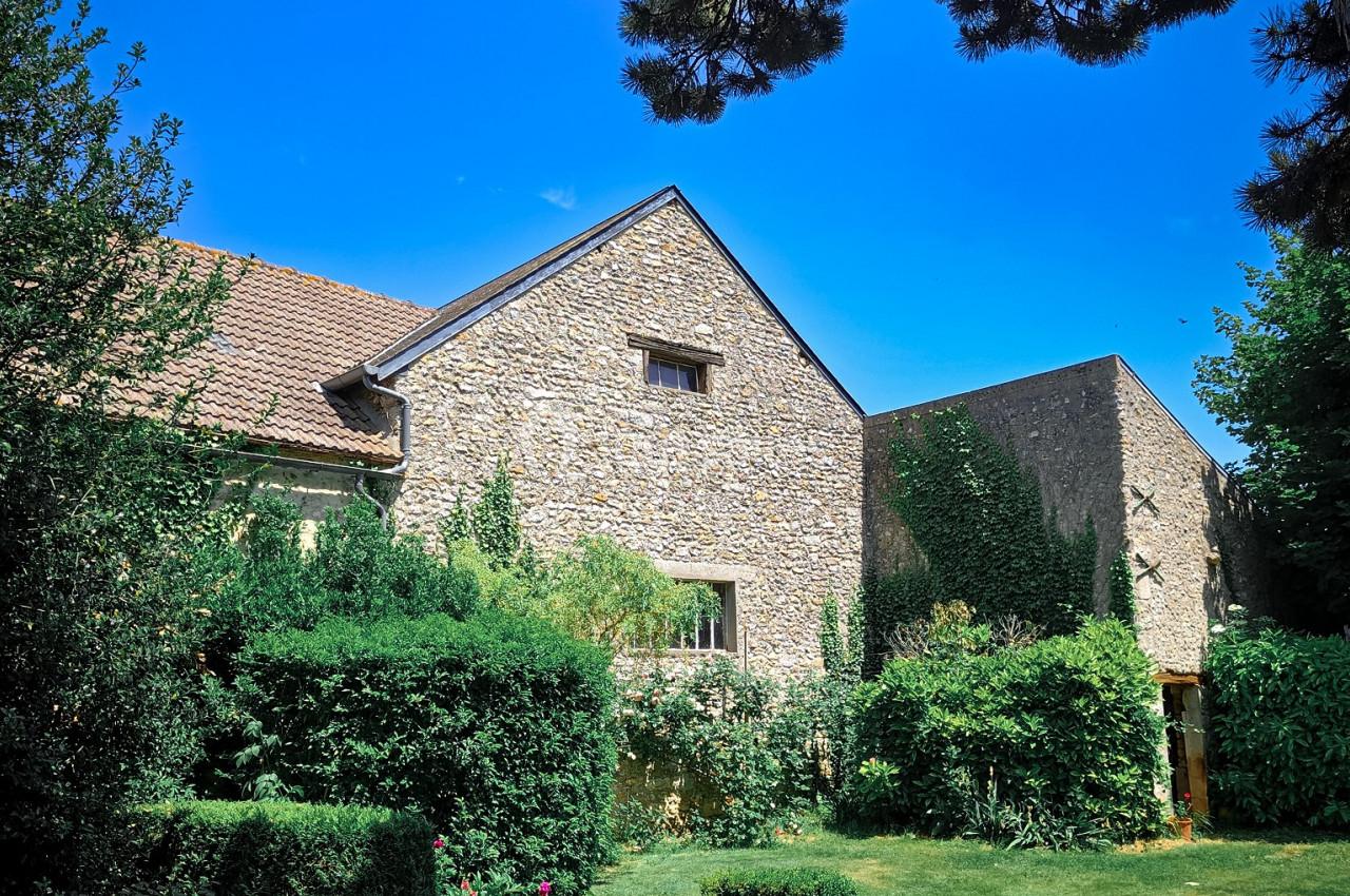 Maison en pierre entourée de verdure avec un jardin arboré sous un ciel bleu clair.