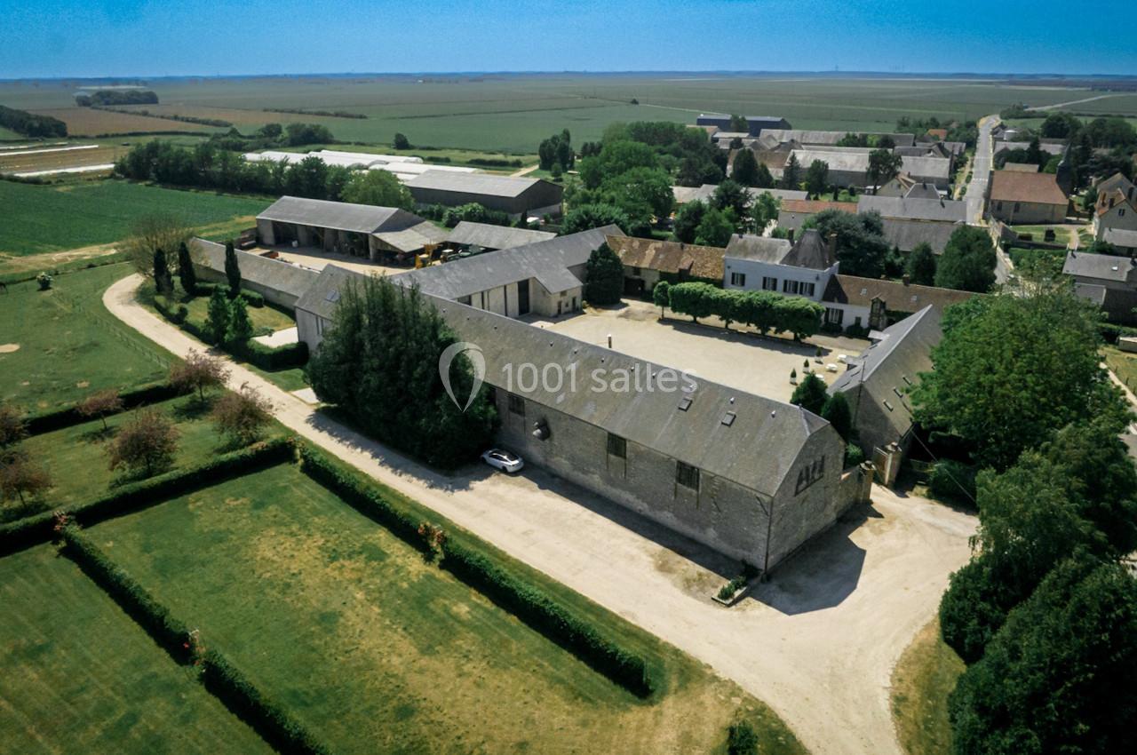 Vue aérienne d'une ferme avec bâtiments en pierre, cour centrale et jardins, entourée de champs et d'un village.