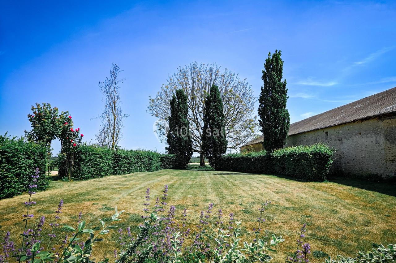 Jardin avec pelouse, haies taillées, arbres et ciel bleu, bordé par un bâtiment en pierre à droite.