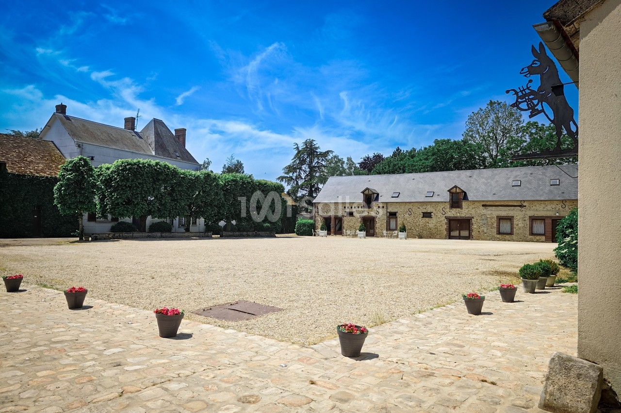 Cour pavée entourée de bâtiments en pierre et végétation, sous un ciel bleu dégagé.