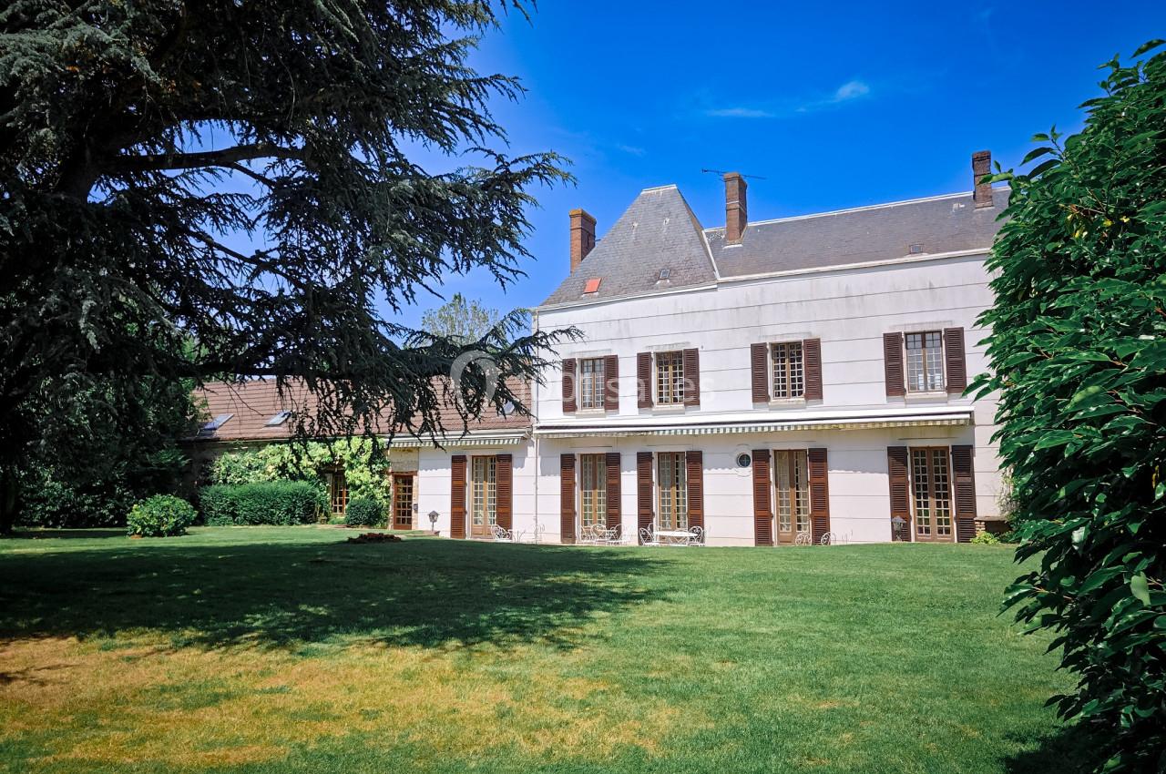 Grande maison avec façade claire, volets marron, entourée d'un jardin verdoyant et d'arbres sous un ciel bleu.